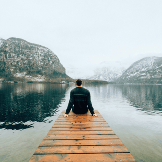 Person sitting on the end of a dock facing a lake and serene mountains